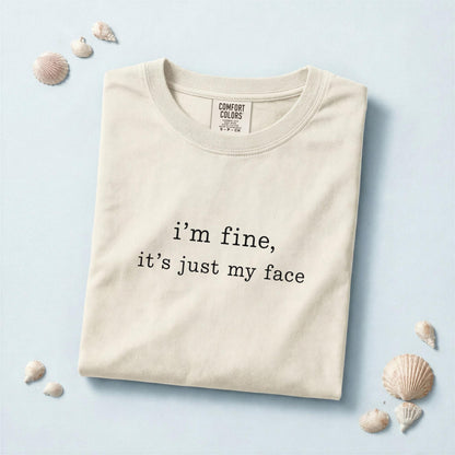 May include: A cream-colored short-sleeve t-shirt featuring the black printed text "i'm fine, it's just my face" centered on the chest. The shirt has a crew neckline and a Comfort Colors brand label visible on the inside of the collar area.