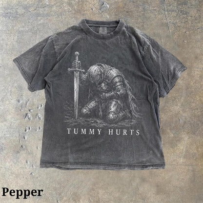 May include: A charcoal gray short-sleeve t-shirt with a crew neckline. The front features a detailed white graphic of a knight in full armor kneeling and holding a sword, with the text "TUMMY HURTS" printed in white capital letters below the knight.