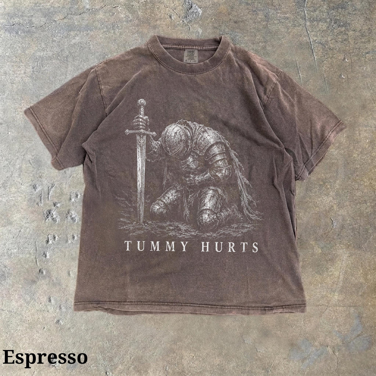 May include: A brown short-sleeve t-shirt featuring a white graphic print of a knight in armor kneeling and holding a sword. Below the graphic, the text "TUMMY HURTS" is printed in white capital letters. The fabric has a faded, washed-out appearance.