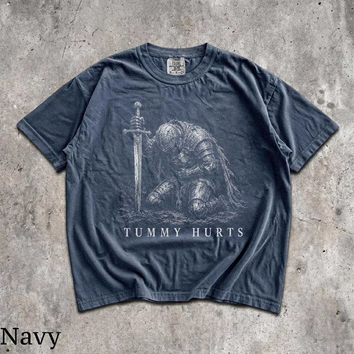 May include: A navy blue short-sleeve t-shirt featuring a white graphic illustration of a knight in full armor kneeling and holding a sword. The text "TUMMY HURTS" is printed in white capital letters below the knight. The shirt has a crew neckline.