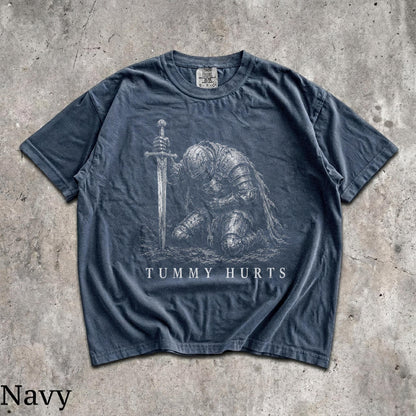 May include: A navy blue short-sleeve t-shirt featuring a white graphic illustration of a knight in full armor kneeling and holding a sword. The text "TUMMY HURTS" is printed in white capital letters below the knight. The shirt has a crew neckline.
