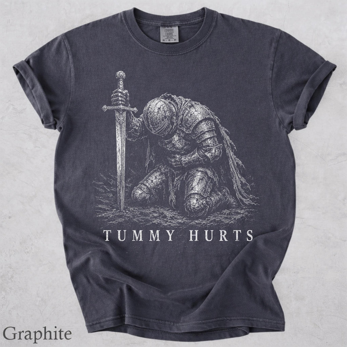May include: A charcoal gray short-sleeved t-shirt featuring a white graphic print of a knight in full armor kneeling and holding a sword upright. The text "TUMMY HURTS" is printed in white capital letters below the knight illustration.