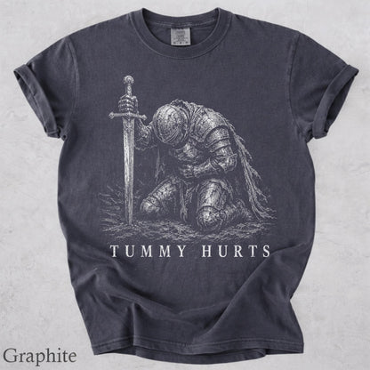 May include: A charcoal gray short-sleeved t-shirt featuring a white graphic print of a knight in full armor kneeling and holding a sword upright. The text "TUMMY HURTS" is printed in white capital letters below the knight illustration.