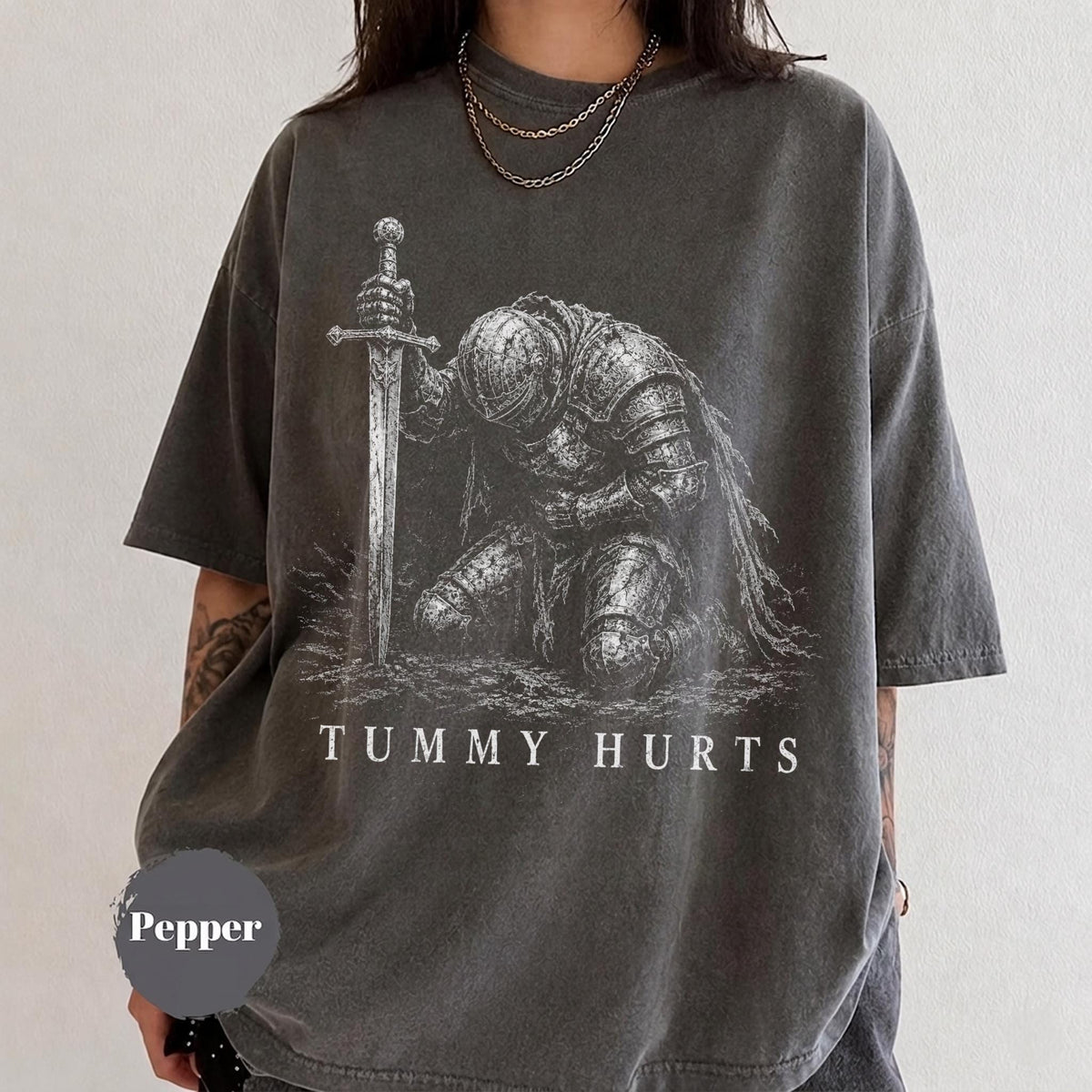 May include: A charcoal gray short-sleeve t-shirt featuring a white graphic print of a knight in armor kneeling and holding a sword. The text "TUMMY HURTS" is printed in white capital letters below the knight. The shirt has a crew neckline.