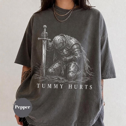 May include: A charcoal gray short-sleeve t-shirt featuring a white graphic print of a knight in armor kneeling and holding a sword. The text "TUMMY HURTS" is printed in white capital letters below the knight. The shirt has a crew neckline.