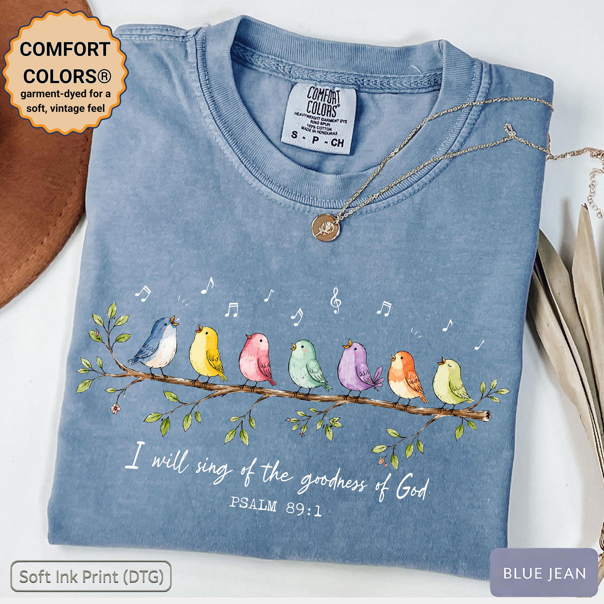 May include: A blue jean colored t-shirt featuring a graphic of seven colorful birds perched on a branch with musical notes. The text reads, I will sing of the goodness of God, PSALM 89:1. The shirt has a Comfort Colors brand label at the neck.