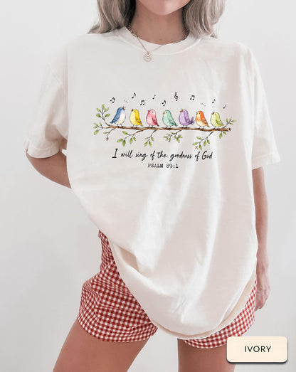 May include: An ivory t-shirt featuring a graphic of seven colorful birds perched on a leafy branch with musical notes above them. Below the branch, the text reads: I will sing of the goodness of God, PSALM 89:1. The shirt has a crew neckline.