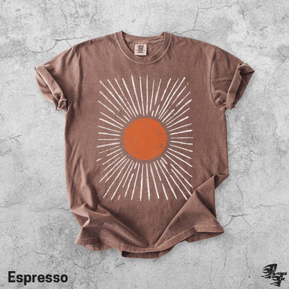 May include: A brown short-sleeve t-shirt featuring a central graphic of a solid orange circle surrounded by radiating white lines. The shirt has a crew neckline and a tag at the inner collar. The fabric has a slightly faded, worn-in appearance.