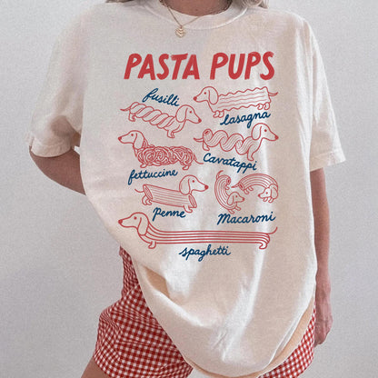 May include: A cream-colored t-shirt featuring the red text PASTA PUPS and red line-art illustrations of dachshunds shaped like various pasta types, labeled in blue cursive text as fusilli, lasagna, fettuccine, cavatappi, penne, macaroni, and spaghetti.