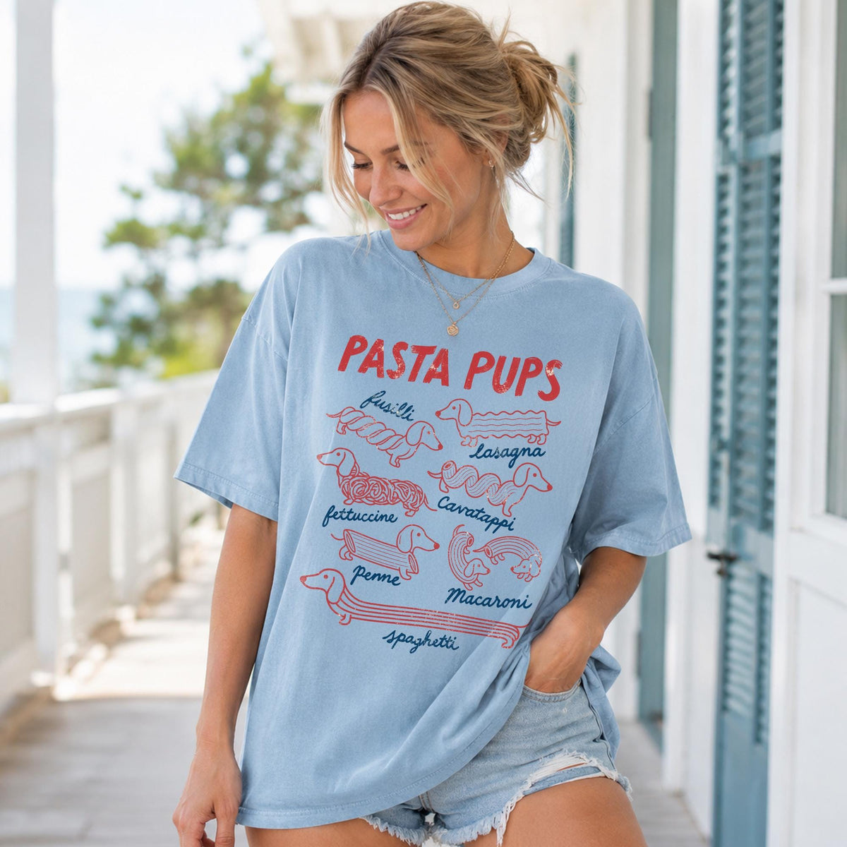 May include: A light blue short-sleeve t-shirt featuring a red and blue graphic print with the text PASTA PUPS. The design displays various dachshund dogs illustrated as different pasta shapes labeled fusilli, lasagna, fettuccine, cavatappi, penne, macaroni, and spaghetti.