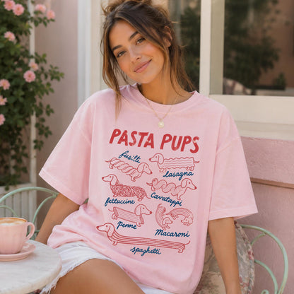May include: A light pink t-shirt featuring the red text PASTA PUPS and various illustrations of dachshunds shaped like pasta types, including fusilli, lasagna, fettuccine, cavatappi, penne, macaroni, and spaghetti, with blue labels for each.