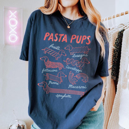 May include: A navy blue short-sleeve t-shirt featuring a red graphic print titled PASTA PUPS. The design displays various dachshund dogs shaped like pasta types labeled fusilli, lasagna, fettuccine, cavatappi, penne, macaroni, and spaghetti.
