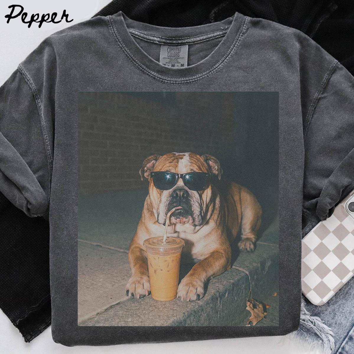May include: A charcoal grey crew neck t-shirt featuring a graphic print of a brown bulldog wearing black sunglasses and drinking from a clear plastic cup with a straw. The word Pepper is printed in black script in the top left corner of the shirt.