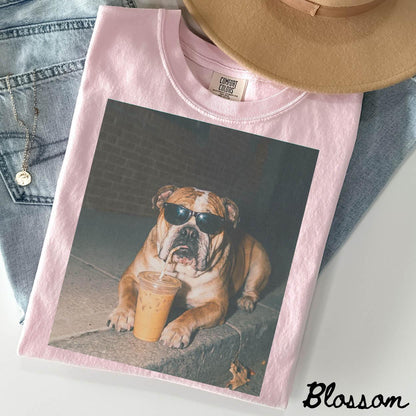 May include: A light pink crewneck t-shirt featuring a graphic print of a brown bulldog wearing black sunglasses and drinking from a clear plastic cup with a straw. The shirt has a Comfort Colors label at the inner neckline and a ribbed collar.