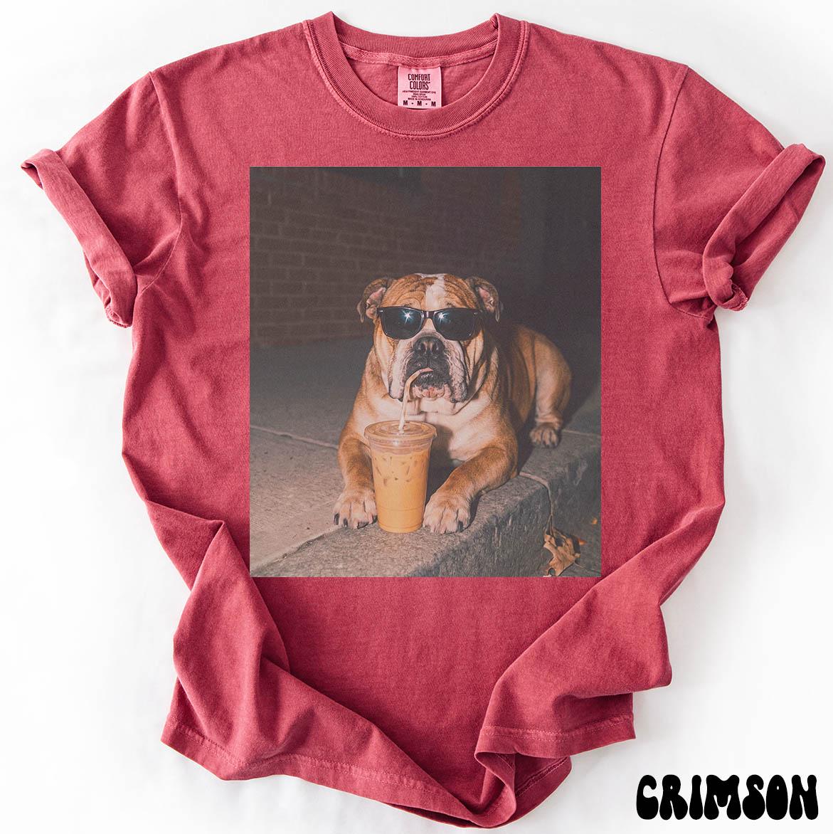 May include: A crimson short-sleeve t-shirt featuring a central graphic print of a tan bulldog wearing black sunglasses and drinking from a clear plastic cup with a straw. The shirt has a crew neckline and a visible brand label at the inner collar.