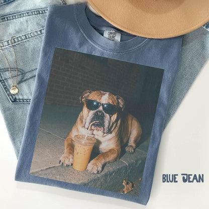 May include: A blue crew neck t-shirt with a Comfort Colors label featuring a graphic print of a brown and white bulldog wearing black sunglasses and drinking from a clear plastic cup with a straw. The shirt is layered with light blue denim fabric.