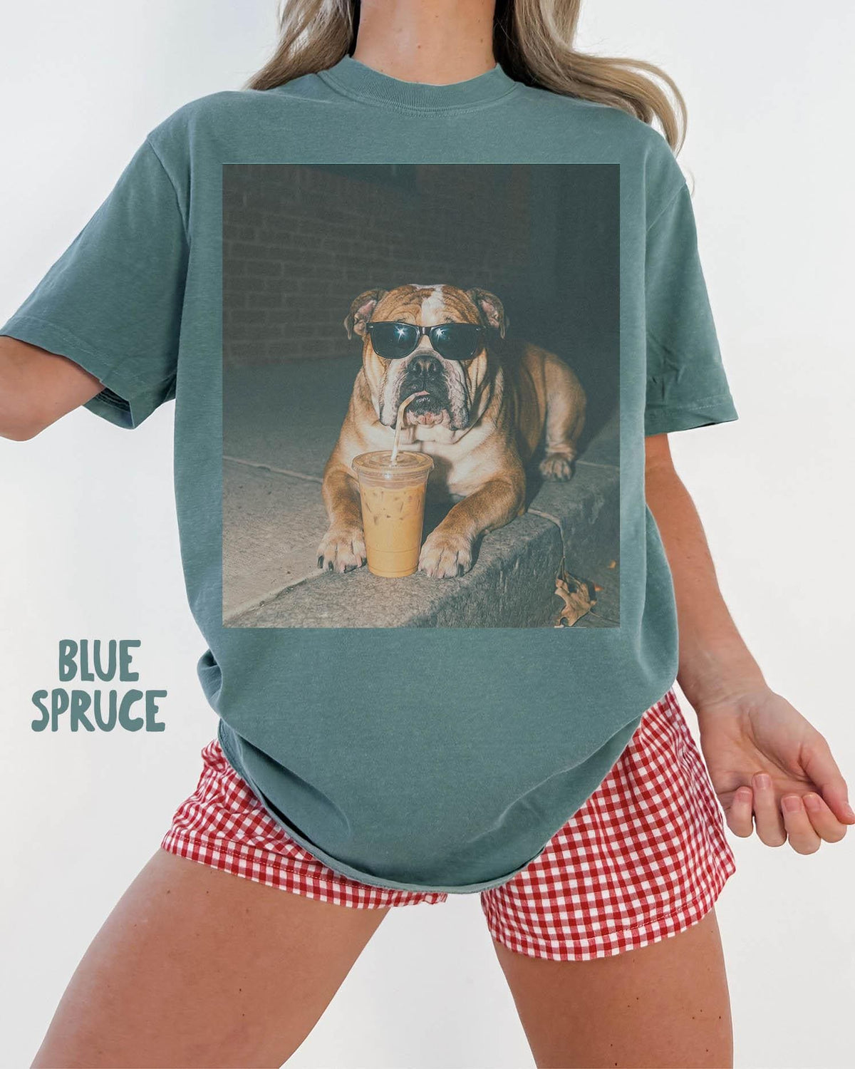 May include: A blue spruce colored t-shirt featuring a graphic print of a brown and white bulldog wearing black sunglasses and drinking from a clear plastic cup with a straw. The text "BLUE SPRUCE" is printed in the lower left corner of the shirt.