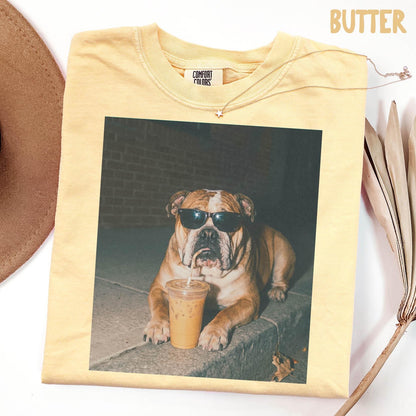 May include: A butter yellow short-sleeve t-shirt featuring a graphic print of a bulldog wearing black sunglasses and drinking from a clear plastic cup with a straw. The shirt has a crew neckline and a Comfort Colors brand label at the inner collar.