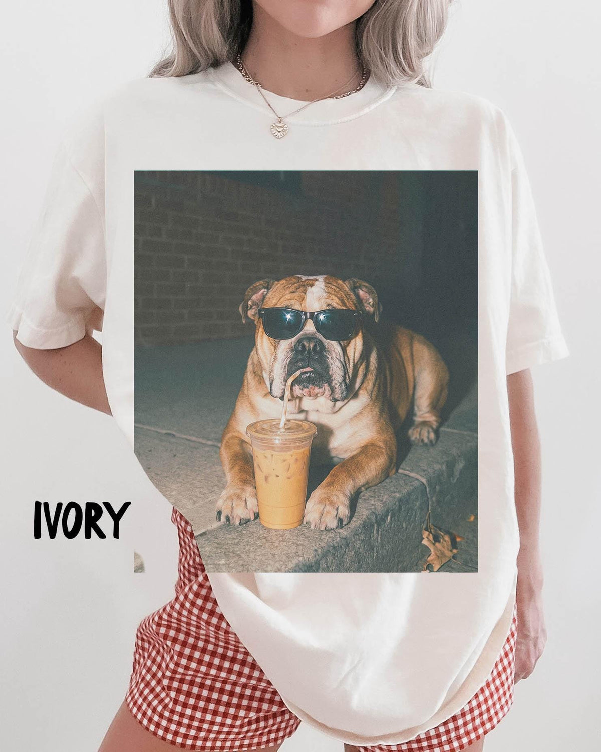 May include: An ivory t-shirt featuring a large graphic print of a brown bulldog wearing black sunglasses and drinking from a clear plastic cup with a straw. The word IVORY is printed in black text on the lower left side of the shirt fabric.