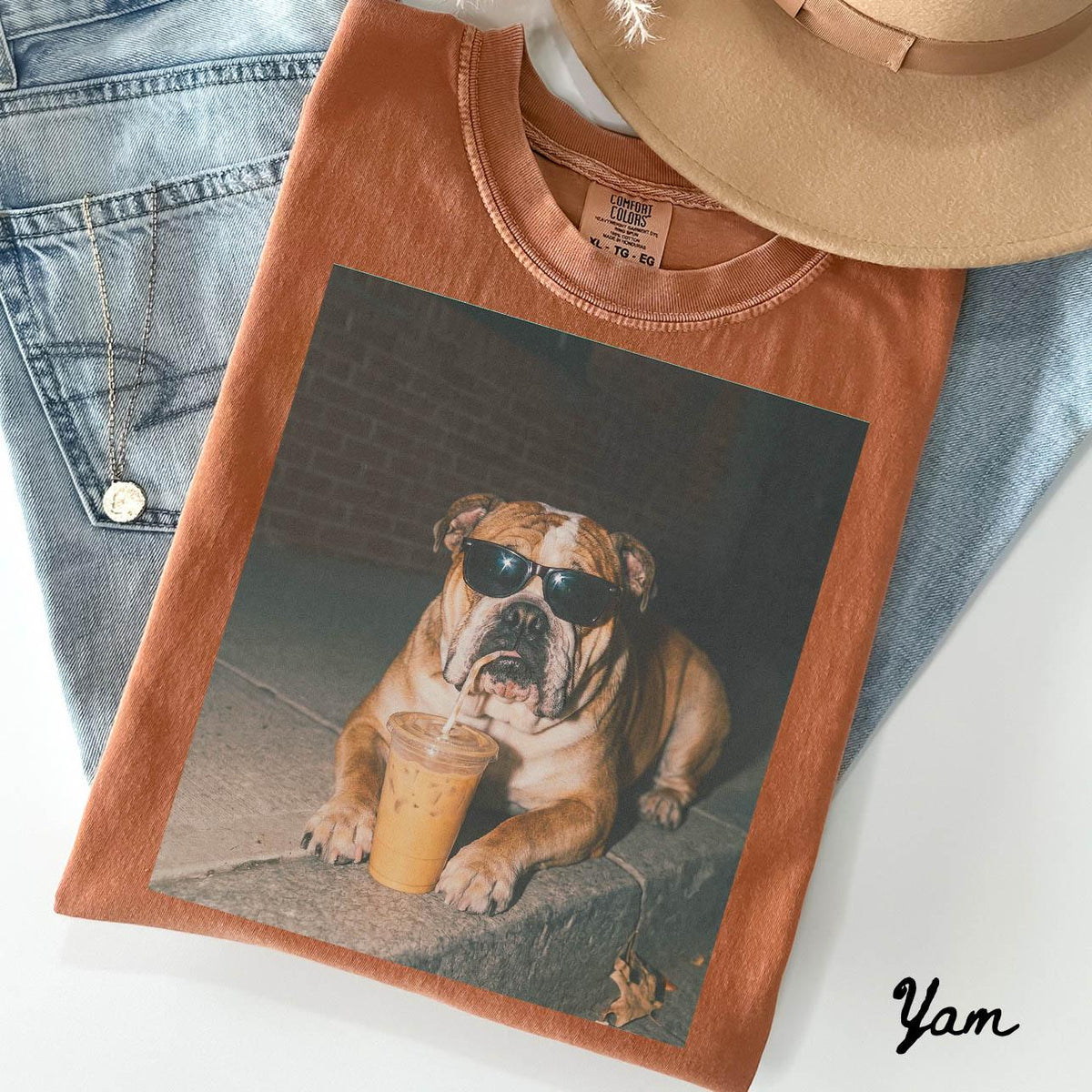 May include: A rust-colored t-shirt featuring a graphic print of a bulldog wearing black sunglasses and holding a clear plastic cup with a straw. The shirt has a crew neckline and a Comfort Colors label visible at the inner collar area.