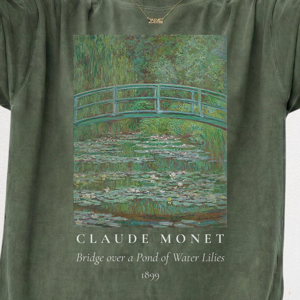May include: A sage green t-shirt featuring a printed reproduction of Claude Monet's painting, Bridge over a Pond of Water Lilies, 1899. The graphic displays a green arched bridge over a pond filled with water lilies, with the text below the image.