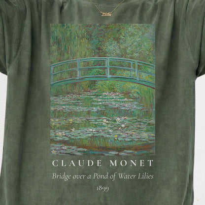 May include: A sage green t-shirt featuring a printed reproduction of Claude Monet's painting, Bridge over a Pond of Water Lilies, 1899. The graphic displays a green arched bridge over a pond filled with water lilies, with the text below the image.