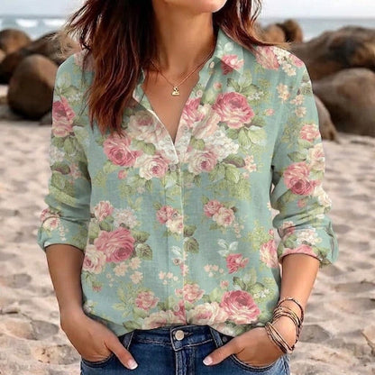 May include: A long-sleeve button-down shirt in a light sage green color featuring an all-over floral print with pink roses and white blossoms. The shirt has a classic collar, a front button placket, and is paired with blue denim jeans.