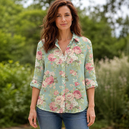 May include: A long-sleeved button-down shirt in a light sage green color featuring a repeating floral pattern of pink and white roses with green leaves. The shirt has a classic collar, a front button placket, and sleeves rolled to the elbows.