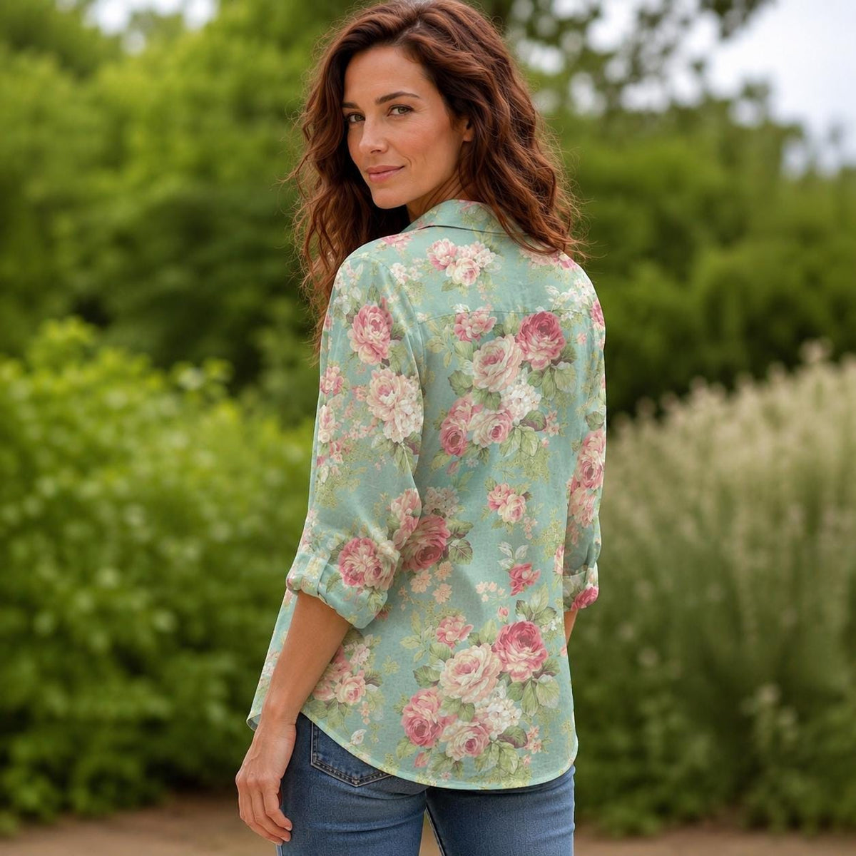 May include: A long-sleeved button-down shirt in a light sage green color featuring an all-over floral print with various shades of pink and white roses. The shirt has a classic collar, a buttoned front, and sleeves that are rolled up to the elbows.