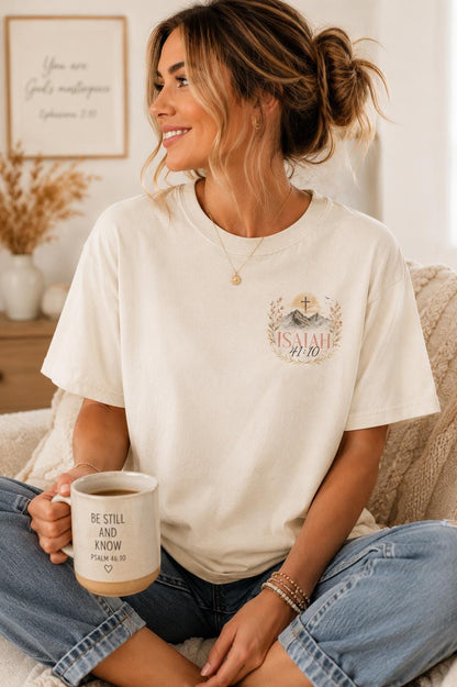 May include: An off-white short-sleeve t-shirt featuring a graphic on the chest with a cross, mountains, and the text Isaiah 41:10. The shirt is paired with a ceramic mug printed with the text Be Still and Know Psalm 46:10 and a heart symbol.