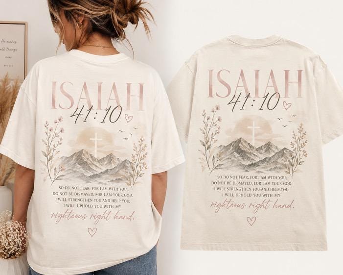 May include: A cream-colored t-shirt featuring a graphic print on the back. The design includes the text "ISAIAH 41:10", a mountain landscape with a cross, floral elements, and the full Bible verse "So do not fear, for I am with you..."