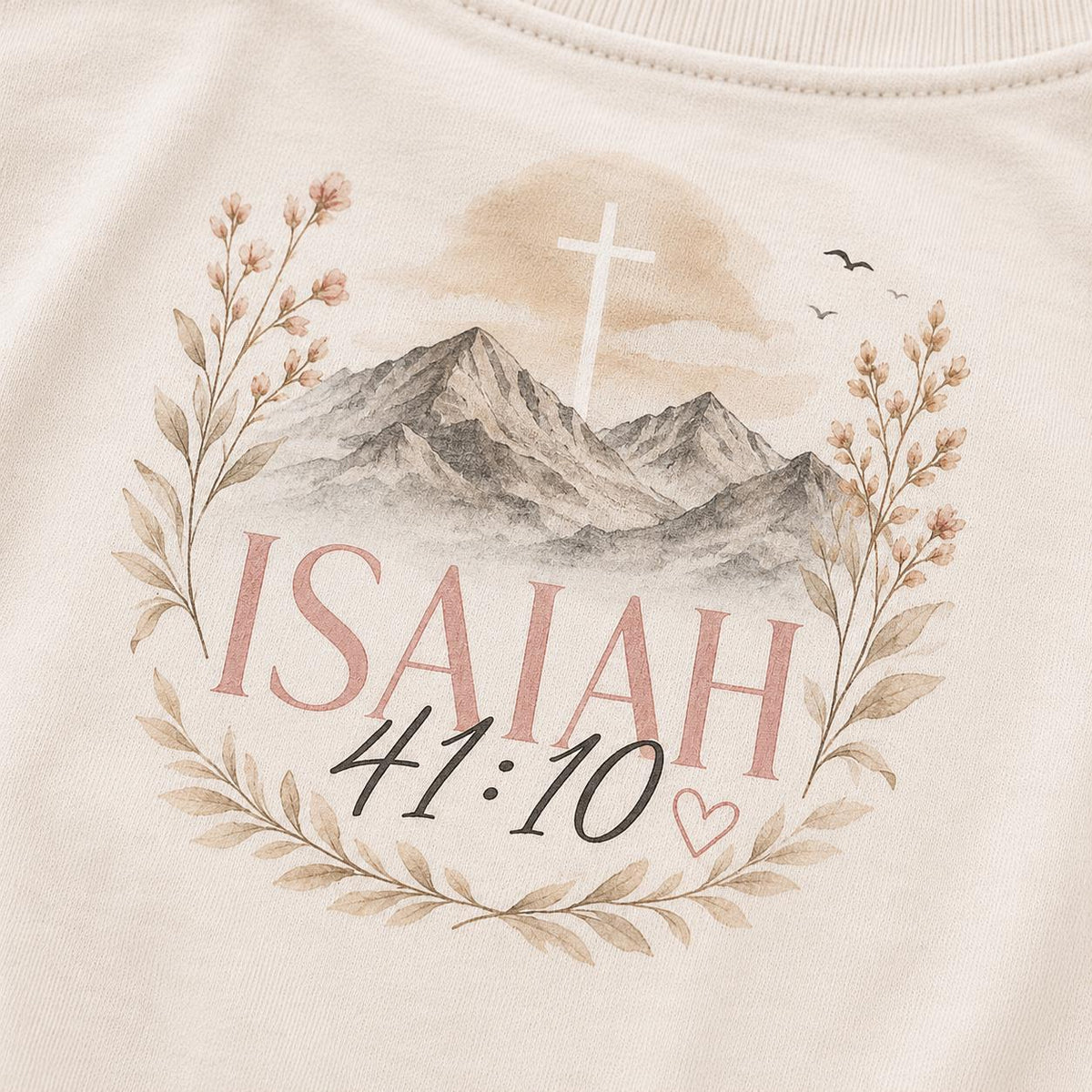 May include: A cream-colored sweatshirt featuring a printed graphic of mountains, a white cross, and floral branches. The text reads ISAIAH 41:10 in pink and black lettering, accompanied by a small heart outline and two birds in the sky.
