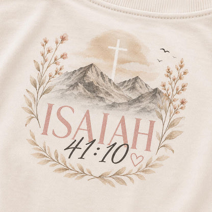 May include: A cream-colored sweatshirt featuring a printed graphic of mountains, a white cross, and floral branches. The text reads ISAIAH 41:10 in pink and black lettering, accompanied by a small heart outline and two birds in the sky.