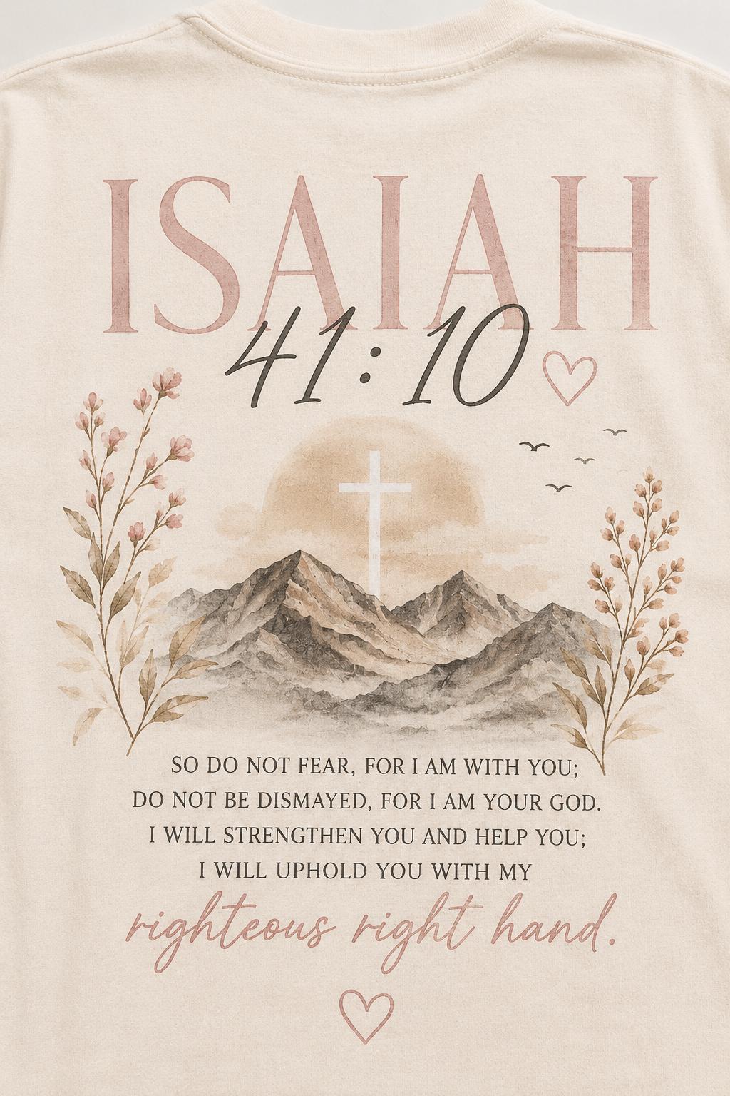 May include: A cream-colored t-shirt featuring a graphic print of mountains, a cross, and floral sprigs. The text reads: ISAIAH 41:10, SO DO NOT FEAR, FOR I AM WITH YOU; DO NOT BE DISMAYED, FOR I AM YOUR GOD. I WILL STRENGTHEN YOU AND HELP YOU; I WILL UPHOLD YOU WITH MY righteous right hand.