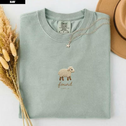 May include: A sage green crewneck t-shirt featuring an embroidered cream-colored sheep with brown hooves and face. Below the sheep, the text reads "found" in cursive and "LUKE 15" in block letters. A silver star pendant necklace rests on the collar.
