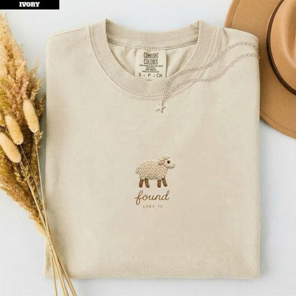 May include: An ivory short-sleeve t-shirt featuring an embroidered design of a small white sheep. Below the sheep, the word "found" is embroidered in a cursive script, followed by the text "LUKE 15" in a smaller, simple font.