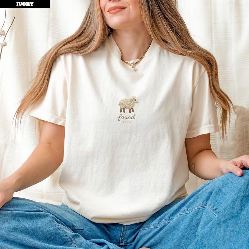 May include: An ivory short-sleeve t-shirt featuring a small embroidered graphic of a fluffy beige sheep centered on the chest. Below the sheep, the word "found" is embroidered in a simple, lowercase brown script font.