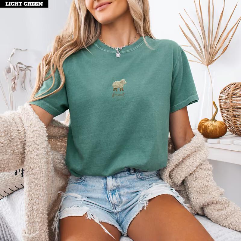 May include: A light green short-sleeved t-shirt featuring a small embroidered graphic of a beige sheep centered on the chest. The shirt has a crew neckline and a relaxed fit, paired with light blue denim shorts with frayed hem details.