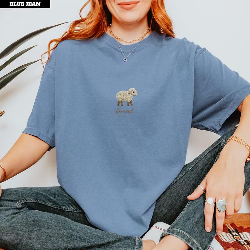 May include: A dusty blue short-sleeve t-shirt featuring a small embroidered graphic of a cream-colored sheep centered on the chest. Below the sheep, the word "found" is embroidered in a simple, lowercase brown script font on the fabric.