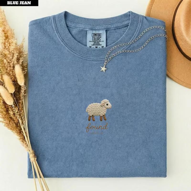 May include: A blue short-sleeve t-shirt featuring an embroidered cream-colored sheep with brown legs and the word "found" stitched in brown thread below it. The shirt has a crew neckline and a Comfort Colors brand label inside the collar.