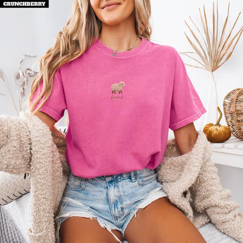 May include: A bright pink short-sleeve t-shirt featuring a small embroidered beige sheep graphic centered on the chest with the word found written in cursive script below it. The shirt has a crew neckline and a relaxed fit.