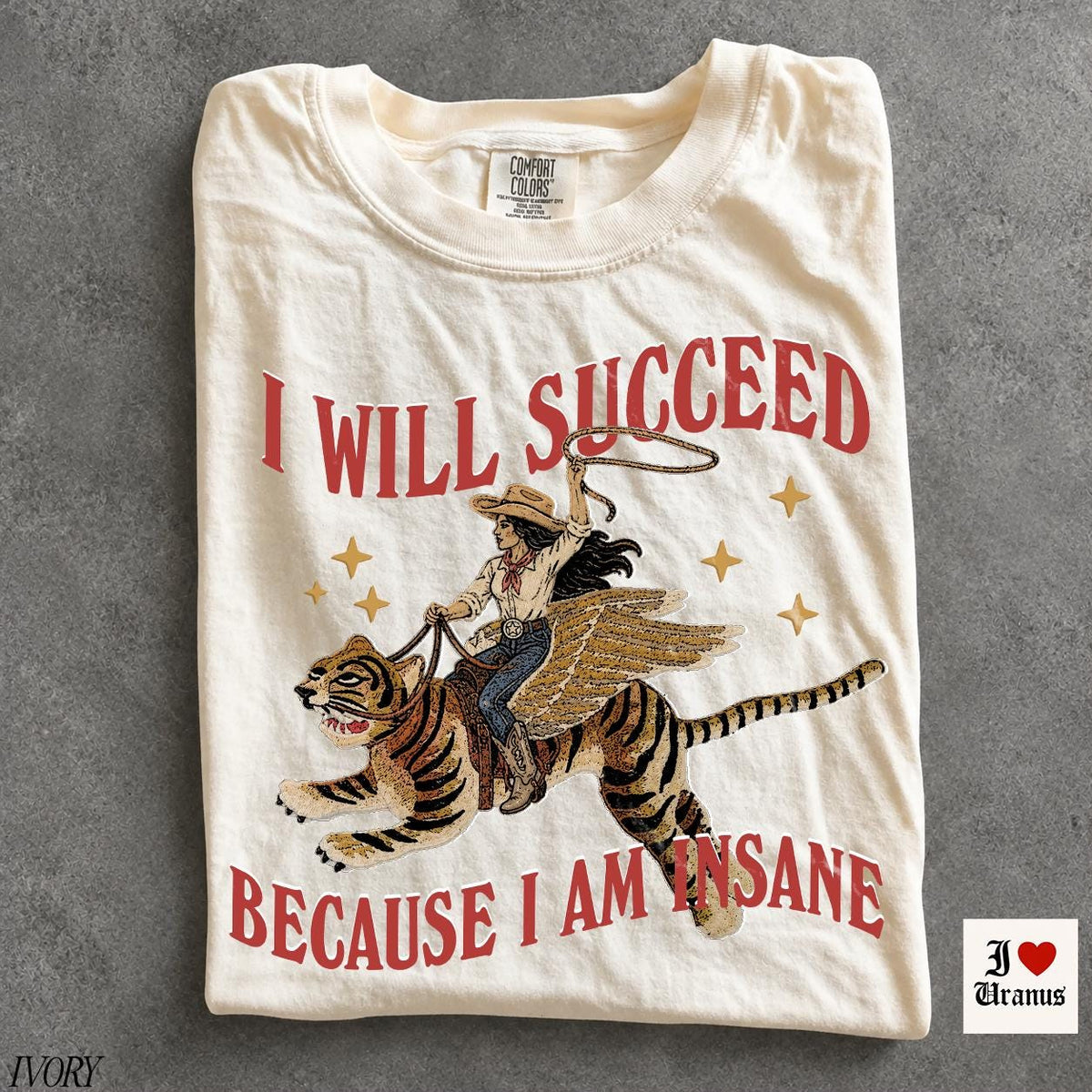 May include: An ivory t-shirt featuring a graphic of a person in a cowboy hat riding a winged tiger while holding a lasso. The text reads I WILL SUCCEED BECAUSE I AM INSANE in red letters. Gold stars surround the central illustration.