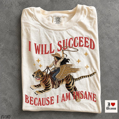 May include: An ivory t-shirt featuring a graphic of a person in a cowboy hat riding a winged tiger while holding a lasso. The text reads I WILL SUCCEED BECAUSE I AM INSANE in red letters. Gold stars surround the central illustration.