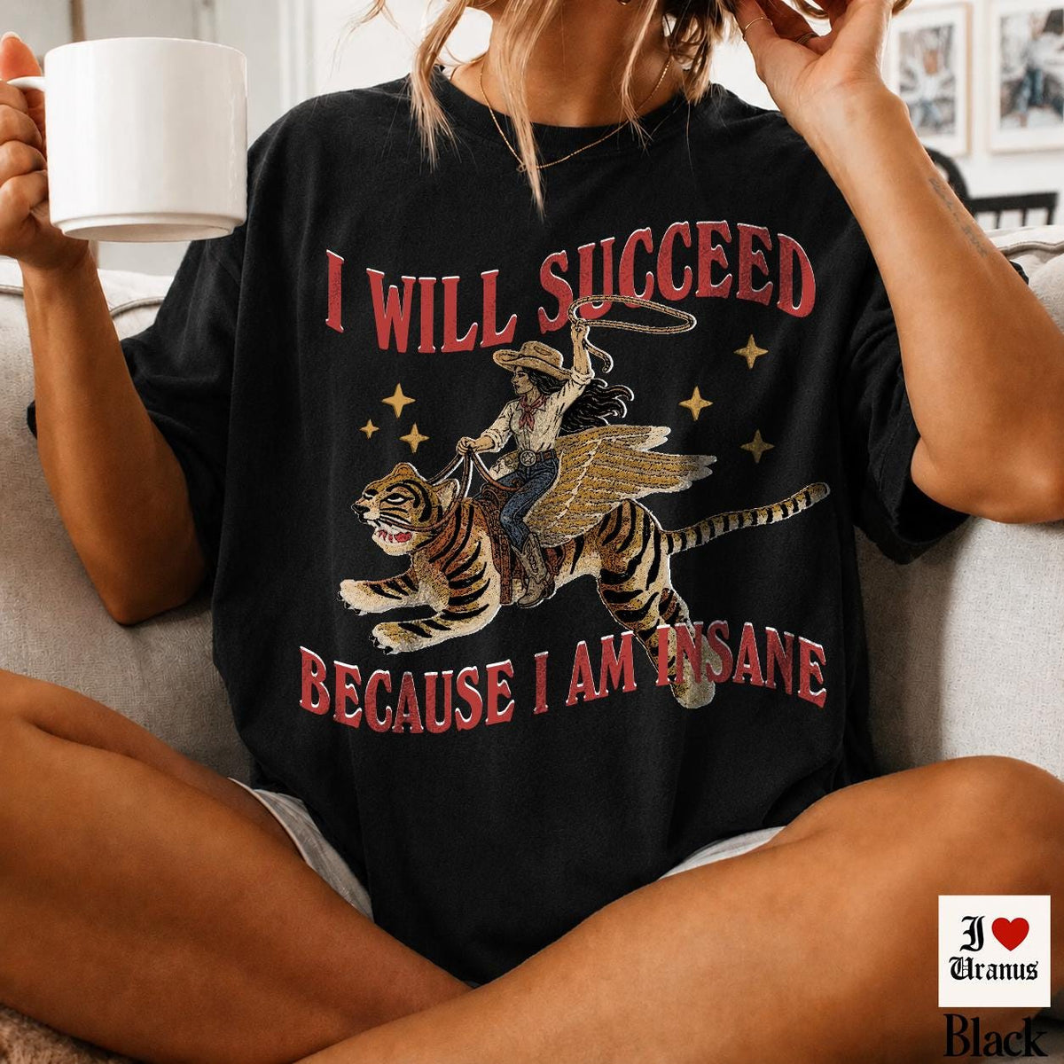 May include: A black short-sleeve t-shirt featuring a graphic of a person riding a winged tiger while holding a lasso. The text "I WILL SUCCEED" appears above the graphic and "BECAUSE I AM INSANE" appears below in red, stylized lettering.