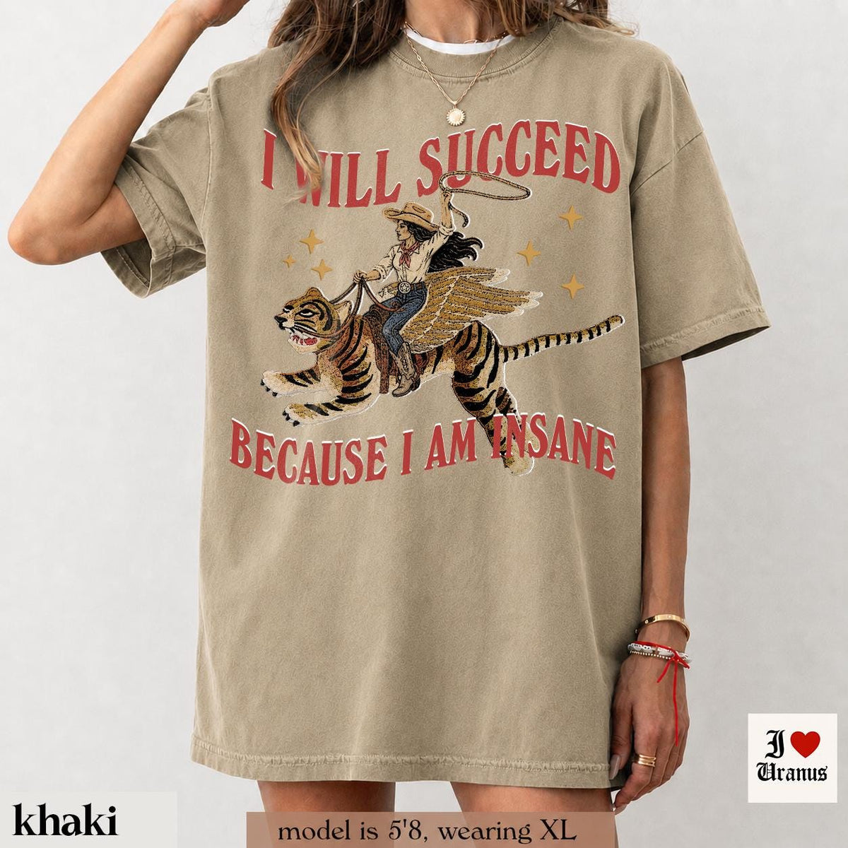 May include: A khaki short-sleeve t-shirt featuring a graphic of a cowgirl riding a winged tiger. The text "I WILL SUCCEED" is printed above the graphic and "BECAUSE I AM INSANE" is printed below in red. Small yellow stars surround the central illustration.