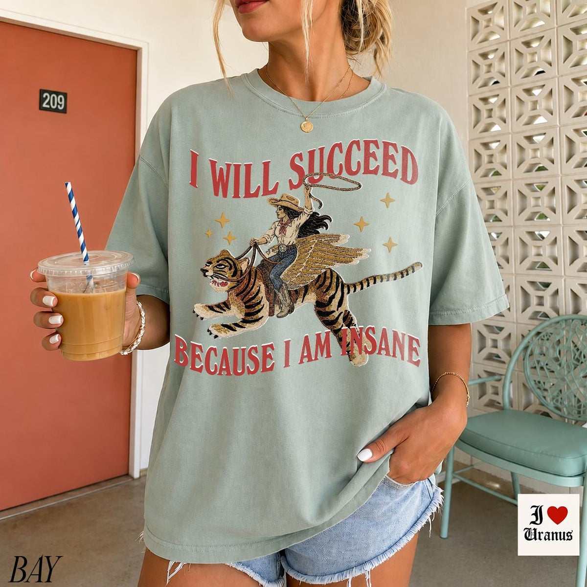 May include: A sage green short-sleeve t-shirt featuring a graphic of a person riding a winged tiger. The text "I WILL SUCCEED BECAUSE I AM INSANE" is printed in red above and below the illustration. The design includes small gold stars.