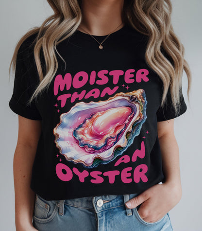 May include: A black short-sleeve t-shirt featuring a colorful graphic of an open oyster shell with pink, blue, and purple hues. The text "MOISTER THAN AN OYSTER" is printed in bold pink letters above and below the oyster illustration.