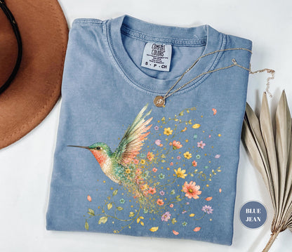 May include: A blue jean colored short-sleeve t-shirt featuring a graphic of a hummingbird with green and orange feathers. The bird appears to be dissolving into a trail of colorful small flowers and leaves. A gold necklace rests on the shirt.