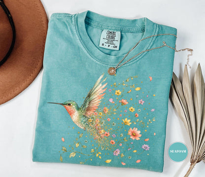 May include: A seafoam green short-sleeve t-shirt featuring a graphic of a hummingbird with wings transitioning into a trail of colorful flowers. The shirt has a crew neckline and a Comfort Colors brand label printed on the inside of the collar.