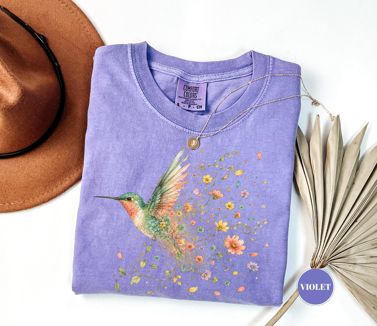 May include: A violet short-sleeve t-shirt with a Comfort Colors label, featuring a graphic of a hummingbird with colorful wings and a trail of small flowers. A thin gold necklace with a small circular pendant rests on the neckline of the shirt.